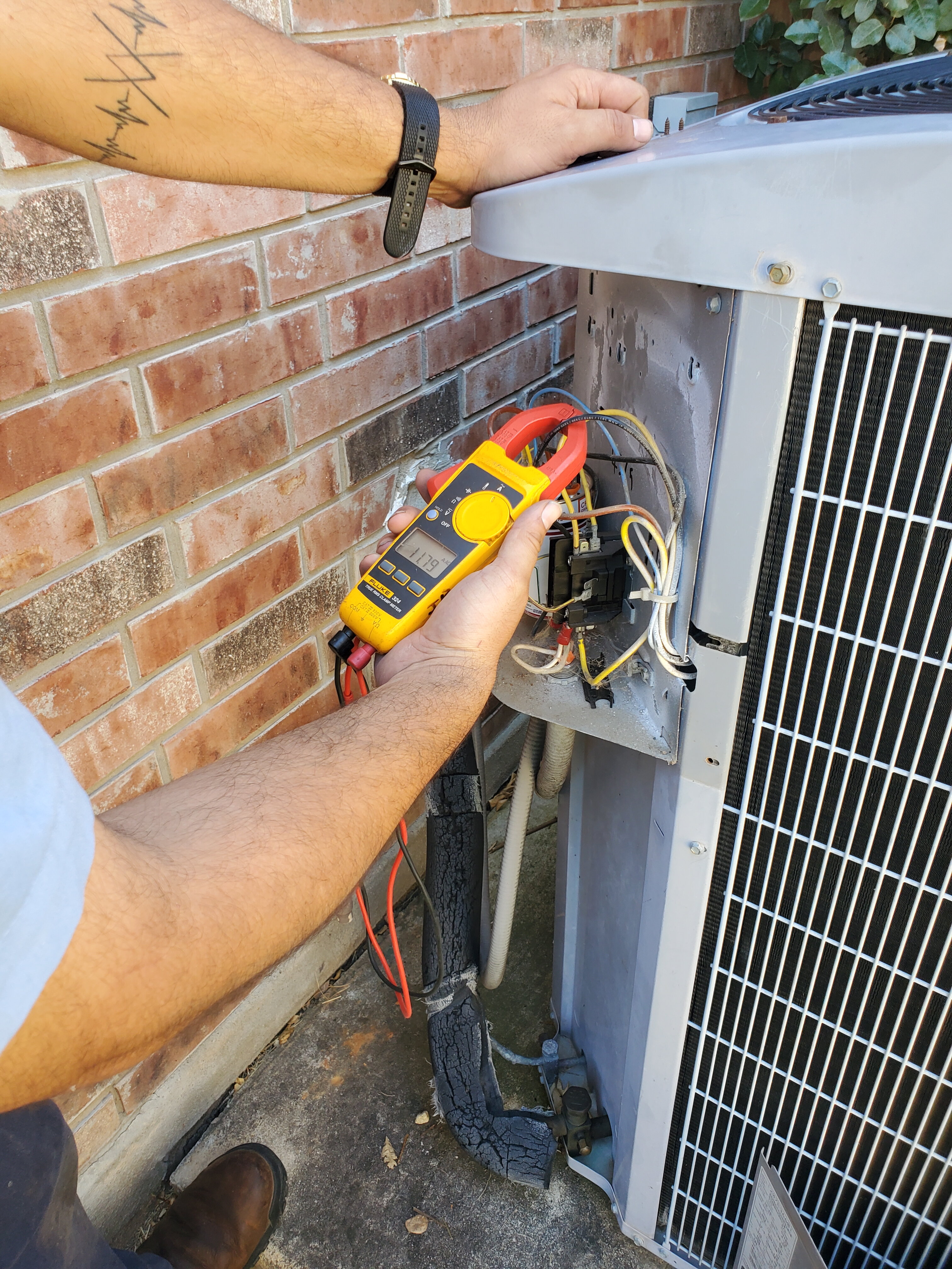Close-up of an AirPro technician using a clamp multimeter to test an open AC condenser unit