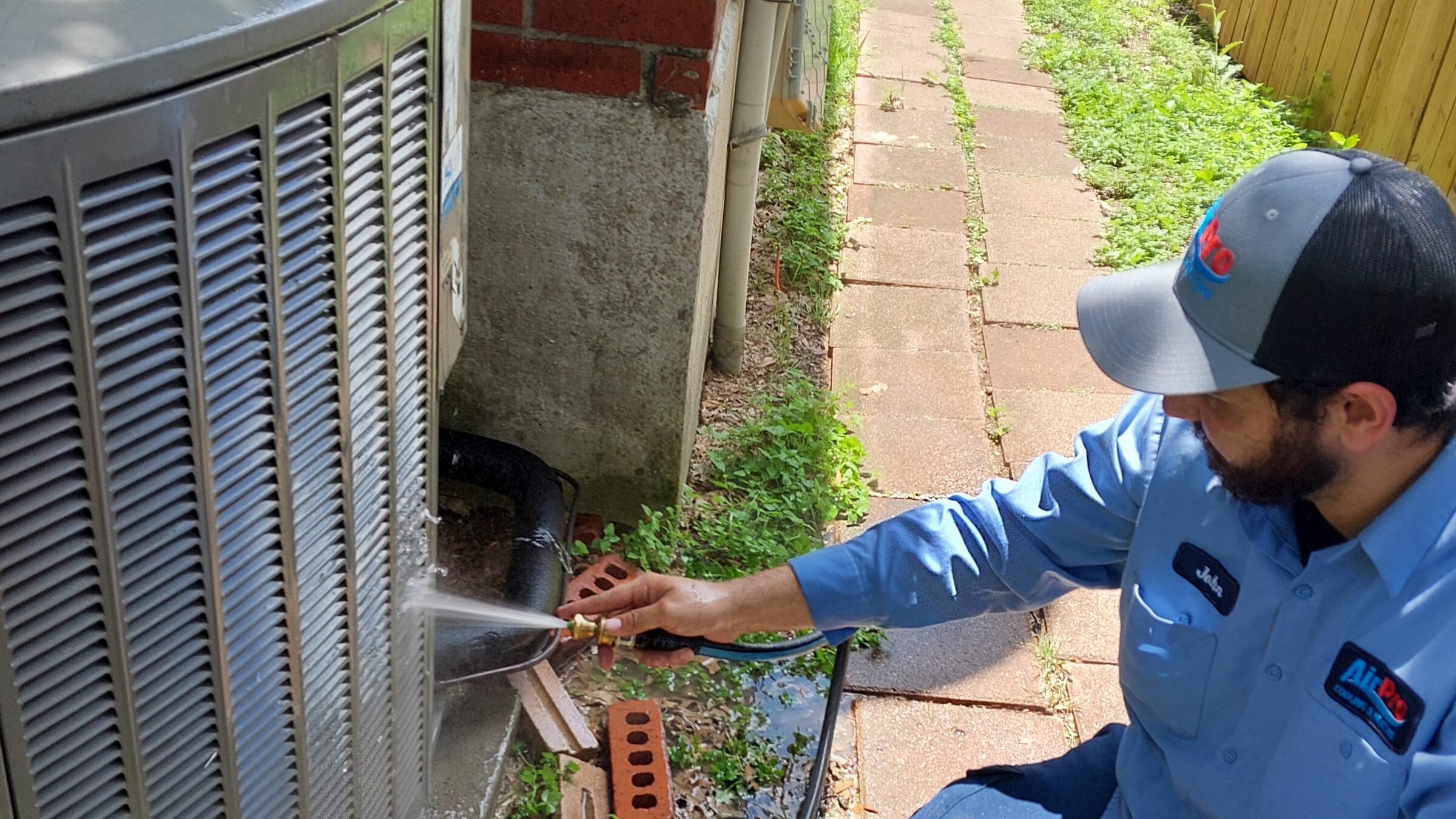 An AirPro technician rinsing the coils on a residential AC condenser during a maintenance visit