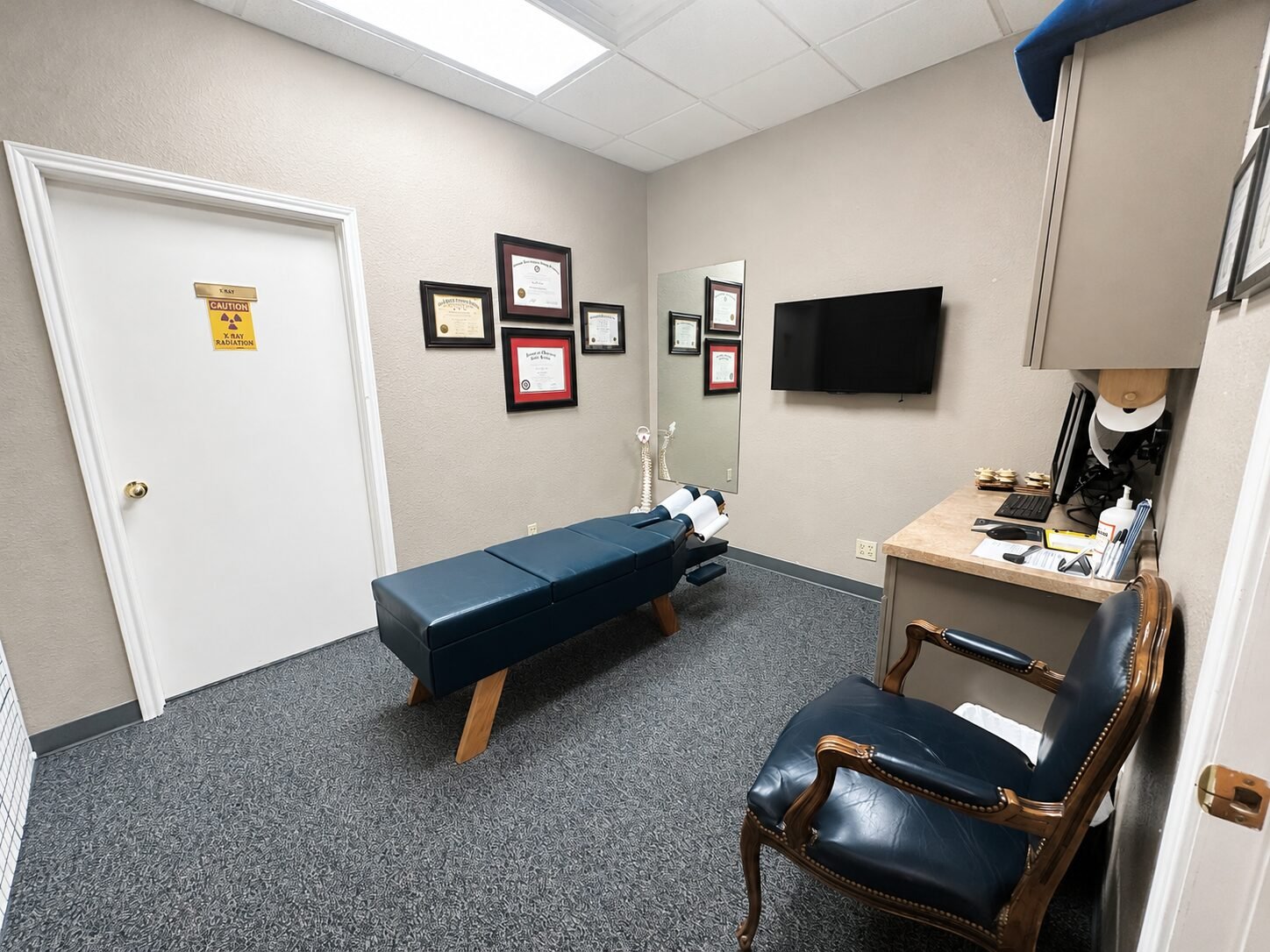 An adjusting room at Bandera Chiropractic, with a navy adjusting table, a spine model, framed certificates on the wall and a door marked for X-ray